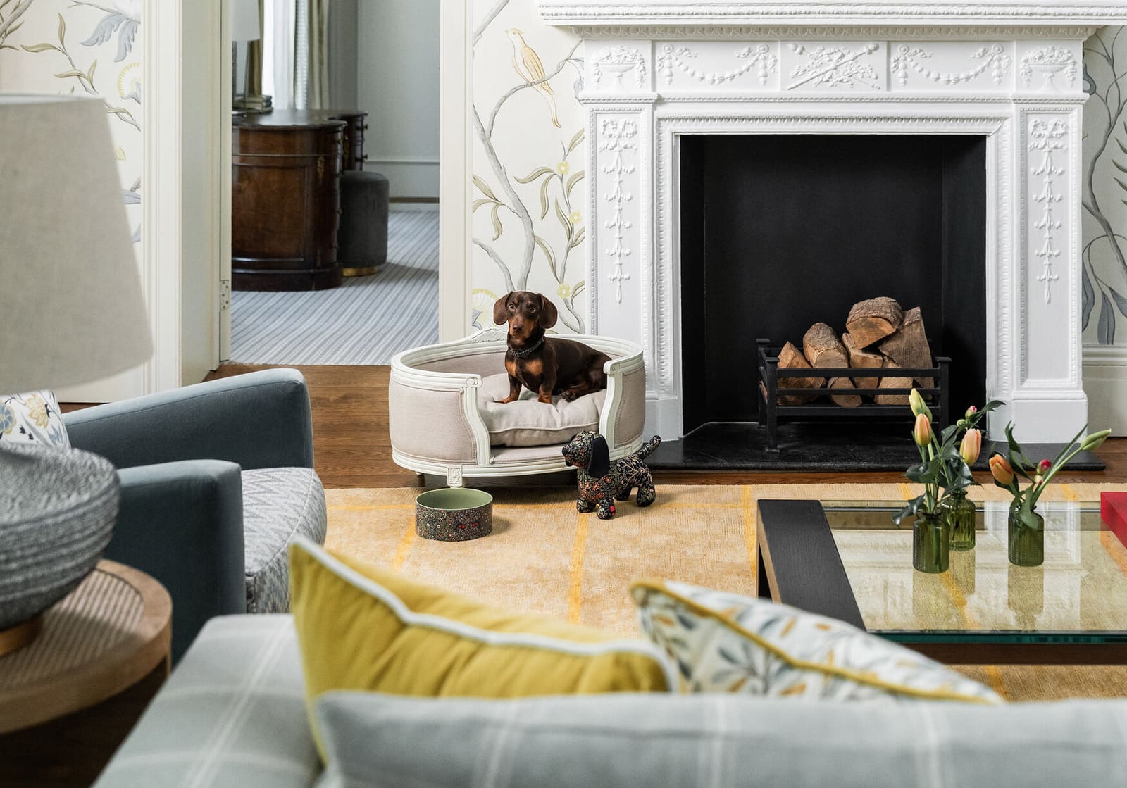 Dachshund sitting up in a bespoke pet bed beside a lit fireplace at Brown's Hotel, Mayfair